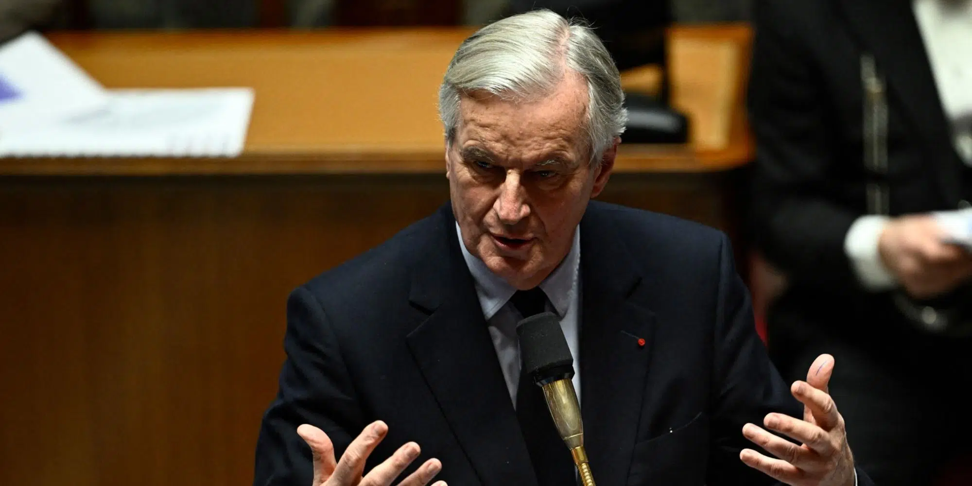 Un homme en costume sombre s’exprime au micro à l’Assemblée nationale, gestes des mains ouverts pour appuyer son discours.
