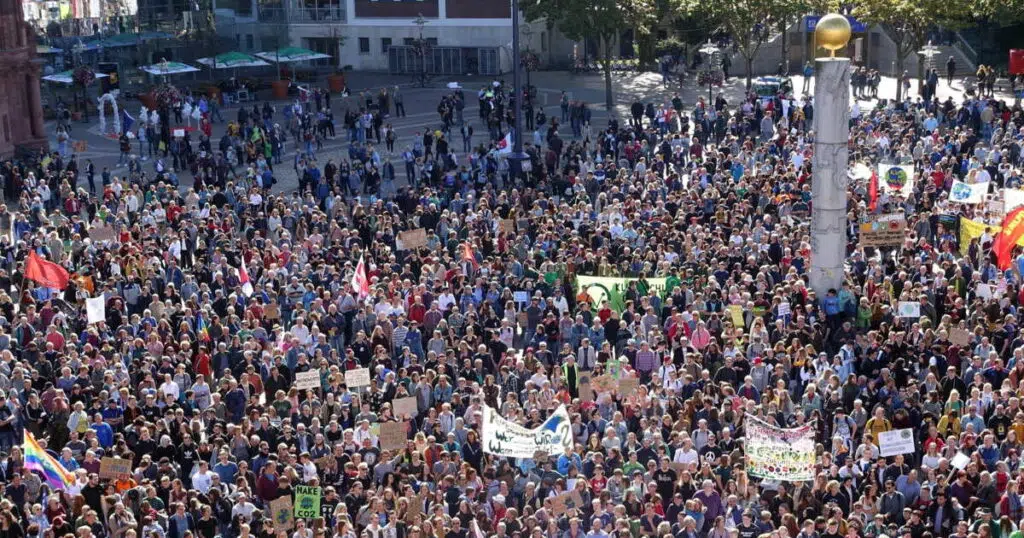 Vue aérienne d’une manifestation de masse en centre-ville, avec une foule dense brandissant pancartes et drapeaux.