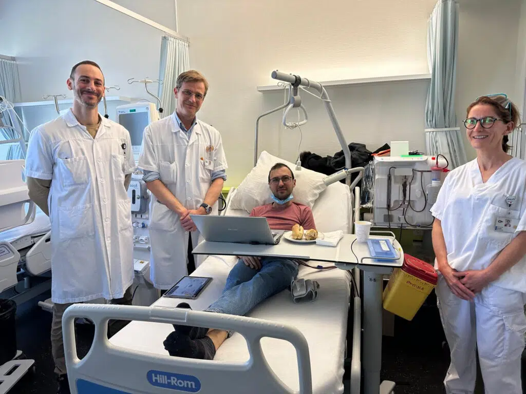 Un patient hospitalisé travaille sur un ordinateur portable depuis son lit, entouré de trois professionnels de santé en blouse blanche dans une chambre d’hôpital moderne.