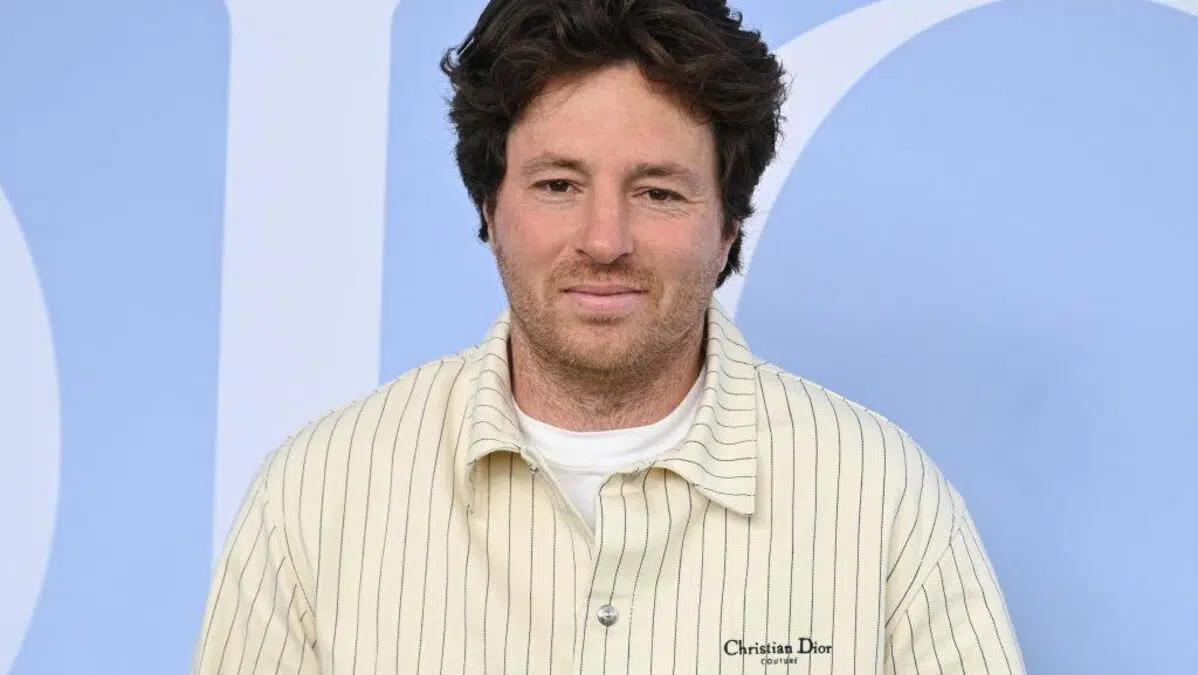 Un homme aux cheveux bruns et bouclés, portant une chemise crème à fines rayures et un T-shirt blanc, pose devant un fond bleu clair.