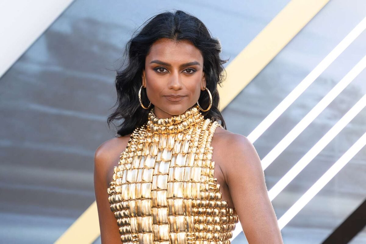 Actrice sur tapis rouge portant une robe dorée et posant devant un décor lumineux lors d’un événement public.