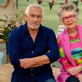 Deux juges assis côte à côte dans un décor de concours de pâtisserie, un homme en chemise bleu marine et une femme en veste rose avec de grandes lunettes rouges.