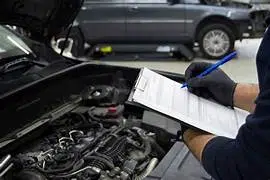 Technicien réalisant un contrôle technique sur une voiture avec le capot ouvert, tenant une fiche de contrôle sur un clipboard dans un garage.