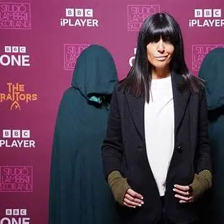 Animatrice de télévision posant devant un mur de logos BBC iPlayer et “The Traitors”, entourée de silhouettes encapuchonnées, lors d’un événement promotionnel.