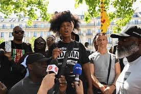 Une femme s’exprime devant des journalistes, entourée de plusieurs personnes lors d’un rassemblement en plein air.