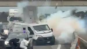 Capture d’une vidéo montrant une explosion près d’un fourgon blindé sur une route italienne, avec des individus en tenue claire à proximité.