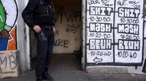 Policier en tenue d’intervention devant l’entrée d’un immeuble, avec des graffitis et des affiches sur le mur.