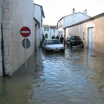 Rue d’un quartier résidentiel inondée : l’eau recouvre la chaussée et entoure des voitures garées devant des maisons.