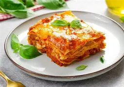 Part de lasagnes maison gratinées servie dans une assiette, avec sauce tomate et feuilles de basilic.
