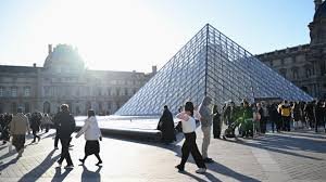 La pyramide du Louvre à Paris, avec des visiteurs qui traversent l’esplanade sous un ciel lumineux.