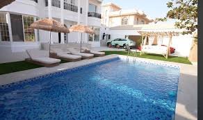 Vue d’une villa moderne avec grande piscine bleue, transats et parasols, dans une cour ensoleillée.