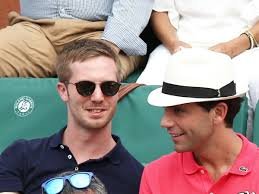 Deux hommes assis côte à côte dans des tribunes : l’un porte des lunettes de soleil et un polo foncé, l’autre un chapeau clair et un polo rouge, ils discutent en souriant.