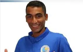 Portrait d’un homme souriant portant un polo bleu avec un écusson, sur fond blanc.