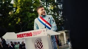 Jeune homme blond portant une écharpe tricolore bleu-blanc-rouge et une chemise claire, debout sur un podium extérieur lors d’un meeting politique, micro devant lui, avec une banderole « MEETING » et un public en arrière-plan sous des arbres.