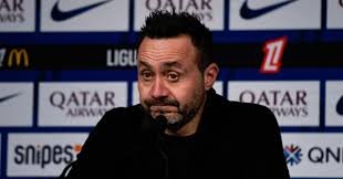 Entraîneur de football assis en conférence de presse, devant un mur de sponsors, l’air sérieux.