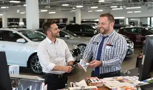 Vendeur en concession automobile discutant avec un client et présentant des documents de vente au milieu des voitures.