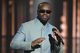 Gims sur scène, lunettes noires et micro en main, lors d’une apparition publique.