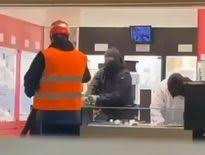 Image d’un braquage dans une bijouterie, avec une personne en gilet orange au premier plan et un individu cagoulé visible à l’intérieur du commerce.