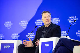 Elon Musk assis sur scène lors d’un forum économique, face à un décor bleu et à des pupitres de conférence.