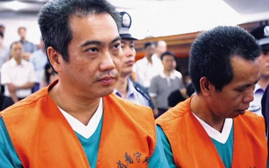 Deux hommes en tenue pénitentiaire orange apparaissent dans une salle d’audience sous la surveillance de policiers