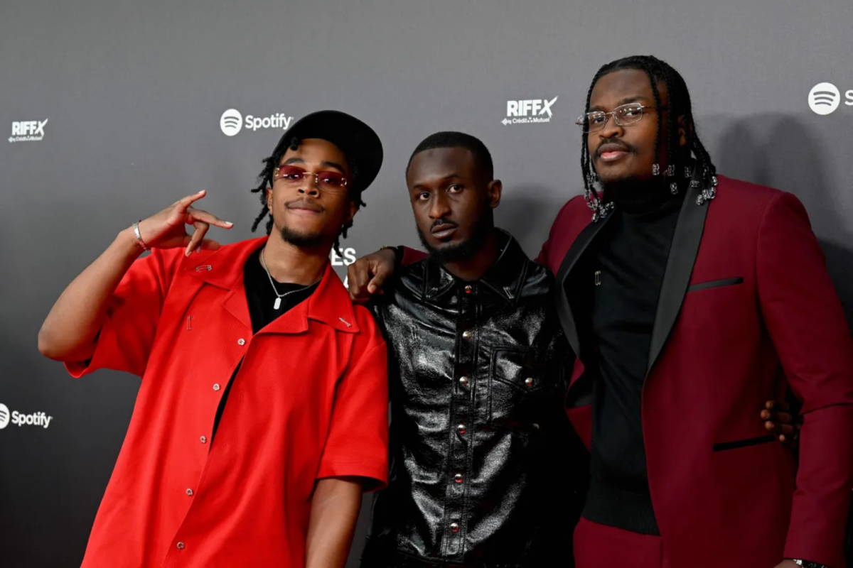 Trois artistes posent sur le tapis rouge des Flammes devant un mur de sponsors, vêtus de tenues noires et rouges.