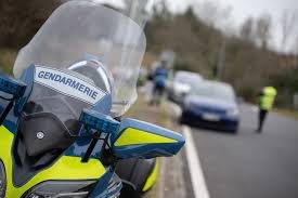 Moto de gendarmerie au bord d’une route, avec des véhicules et des gendarmes visibles en arrière-plan.