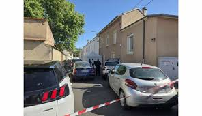 Rue résidentielle bouclée par un ruban de sécurité, avec plusieurs véhicules stationnés et des forces de l’ordre sur place.