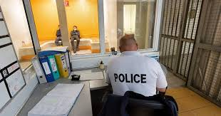 Vue intérieure d’un commissariat avec un policier assis de dos face à deux personnes visibles dans une pièce vitrée.