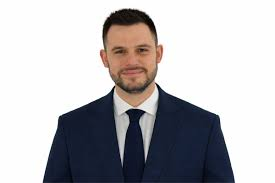 Portrait d’un homme en costume bleu, chemise blanche et cravate sombre, sur fond clair.