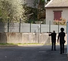 Deux silhouettes dans une rue résidentielle, dont une personne tenant une carabine en direction d’un espace extérieur clos.