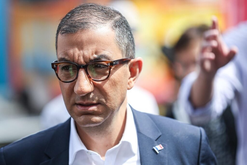 Portrait de Jean Messiha en costume bleu marine et chemise blanche, portant des lunettes, lors d’un déplacement public en extérieur.