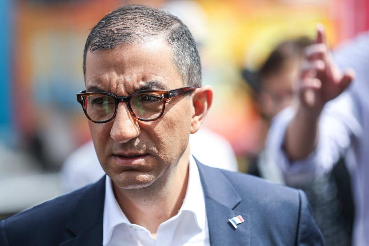 Portrait de Jean Messiha en costume bleu marine et chemise blanche, portant des lunettes, lors d’un déplacement public en extérieur.