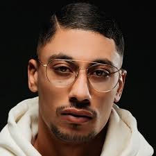 Portrait du rappeur français Maes portant des lunettes transparentes et un sweat à capuche blanc sur fond sombre.