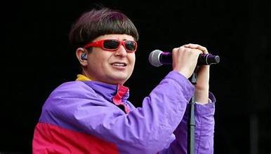 Chanteur américain sur scène, lunettes rouges et veste colorée, tenant un micro devant le public.