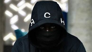 Rappeur cagoulé portant une casquette noire, photographié dans une ambiance sombre et urbaine.