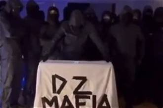 Plusieurs individus cagoulés posent dans l’obscurité derrière une bannière portant l’inscription « DZ Mafia ».